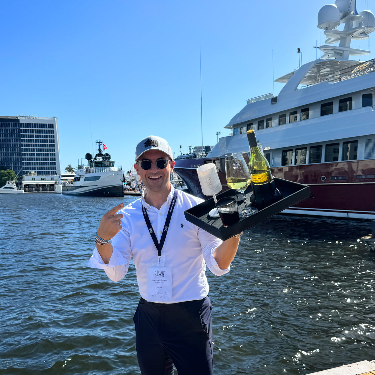 Founder and CEO Christoph with a metal tray in his hand. On it is a magnetic crystal glass WINE, a magnetic plastic glass wine CHEERS in white, a Magnetic drinking cup Triple classic and a bottle of wine. A marina with ships can be seen in the background