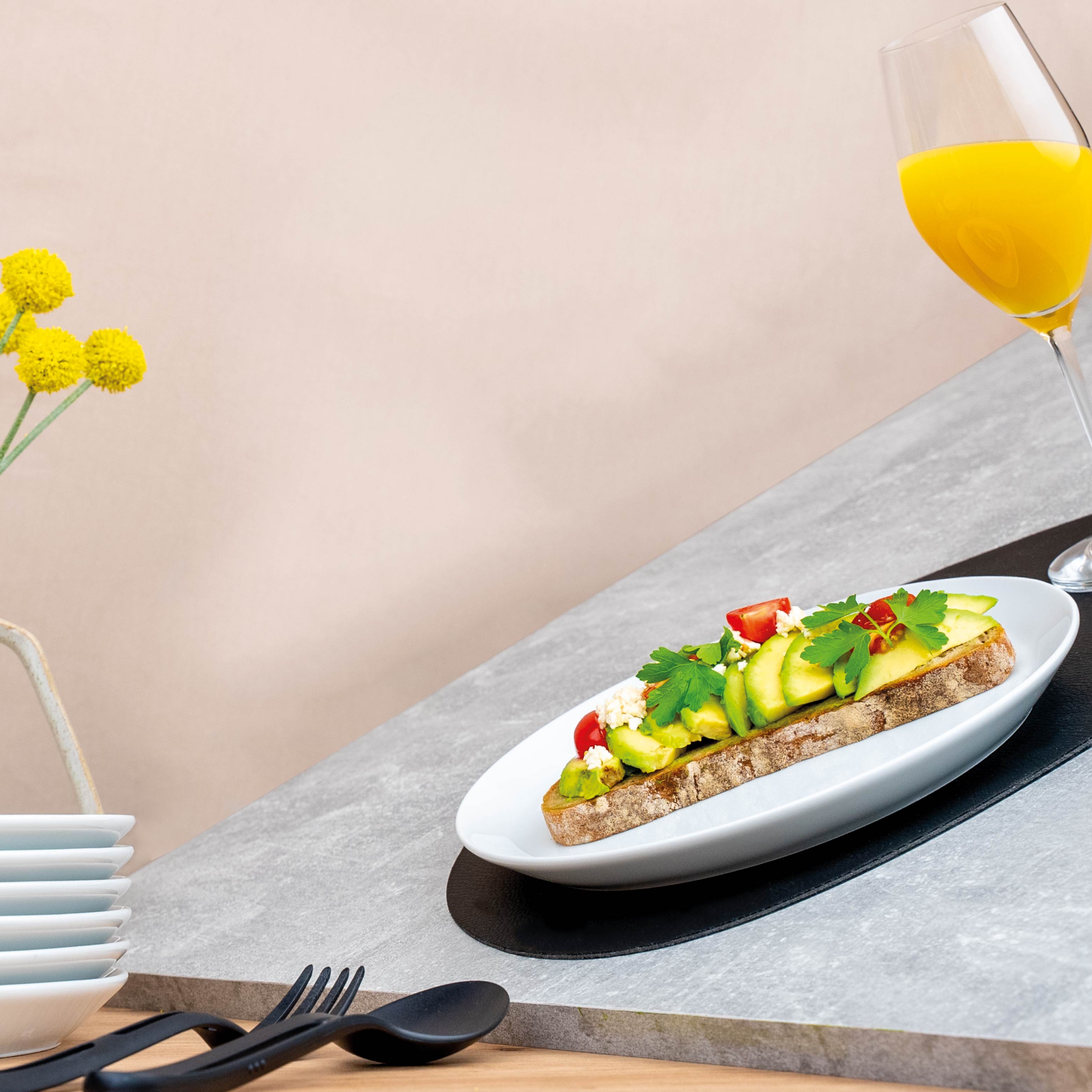Platos de porcelana magnéticos con pan y aguacate por encima y vaso champán relleno de zumo de naranja sobre mantel y Cubiertos negros 